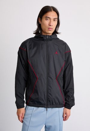Man wearing a black zip-up jacket with red trim and logo, paired with light blue pants, standing against a plain light background.