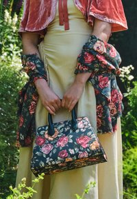 Borsa floreale realizzata in materiale testurizzato nero, con rose rosa e arancioni. Doppie maniglie nere e forma rettangolare. Ambientazione all'aperto popolata.