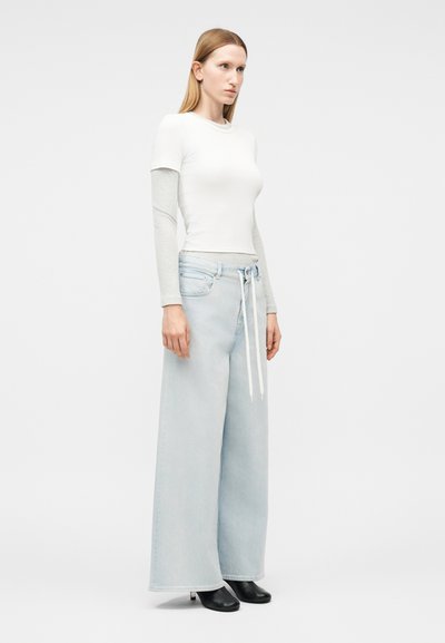 Γυναίκα που στέκεται, φοράει στρωματοποιημένο λευκό και γκρι μακρυμάνικο τοπ, ανοιχτό μπλε τζιν με φαρδιά μπατζάκια και λευκό κορδόνι, και μαύρα μποτάκια στο αστράγαλο.