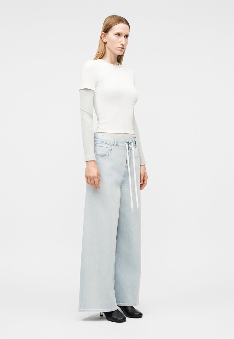 Woman standing, wearing layered white and gray long-sleeve top, light blue wide-leg jeans with white drawstring, and black ankle boots.