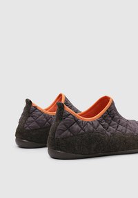 Zapatillas de casa acolchadas de color marrón oscuro con un forro de tela naranja, perfil bajo, textura suave y suelas de goma para mayor agarre. Presentan un acabado redondeado en la parte trasera.