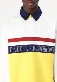 Hvid polo skjorte med en marineblå krave, gul krop og vandrette striber i rød, hvid og blå. Har "HILFIGER" skrevet med store bogstaver.