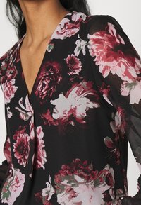 Bloes met bloemenprint op een zwarte achtergrond, doorschijnende lange mouwen en een diepe V-hals. Het ontwerp bevat grote, levendige roze en bordeauxrode bloemen.