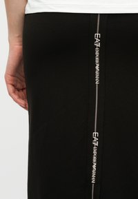 Κοντινό πλάνο μιας μαύρης φούστας με κάθετη λωρίδα λογότυπου EA7 Emporio Armani; μέρος ενός χεριού και λευκό τοπ ορατό.