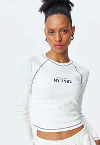 Weißes, geripptes Langarmoberteil mit marineblauen Nähten und "NY 1982" Grafik. Zugeknöpftes Design, taillierte Silhouette und runder Kragen.