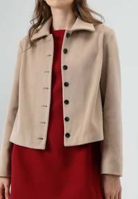 Beige fleecejack met een kraag, voorzien van zes zwarte knopen en een rechte pasvorm. Draagt over een rode jurk. Zachte textuur, minimalistisch ontwerp.