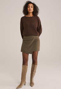 Maglione marrone morbido, gonna avvolgente verde oliva e stivali beige in suede al ginocchio. Il modello posa con le braccia rilassate, evidenti il contrasto di texture e colore.