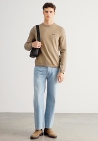 Jasnobrązowy sweter z okrągłym dekoltem i logo, w połączeniu z jasnoniebieskimi jeansami o prostych nogawkach. Brązowe zamszowe botki do kostki i czarna torba.