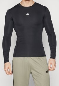 Mand iført tætsiddende langærmet sort træningstop og løse lysegrå Adidas-bukser, der står foran en ensfarvet baggrund.