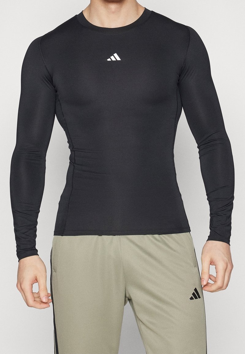 Mand iført tætsiddende langærmet sort træningstop og løse lysegrå Adidas-bukser, der står foran en ensfarvet baggrund.