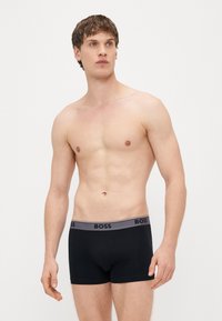 Μαύρα μπόξερ σορτς με γκρι ζωνάρι μέσης που φέρει το λογότυπο "BOSS". Το ύφασμα φαίνεται λείο με εφαρμοστή σχεδίαση. Το μοντέλο δεν φορά πουκάμισο.