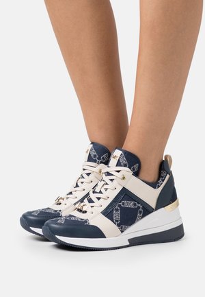 Osoba nosząca granatowe i beżowe wysokie sneakersy z białymi sznurowadłami, warstwową podeszwą i wzorem logo na białym tle.