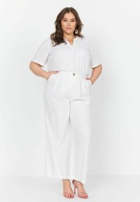 Hvid linned jumpsuit med korte ærmer, knaplukning og sidelommer. Har brede ben og en afslappet pasform. Bæres med lyserøde sandaler.