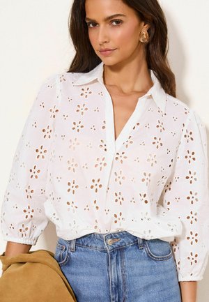 Nő fehér, virágmintás lyukacsos blúzban, puffos ujjakkal és kék farmerral, kézben egy barna velúr dzsekit tart.