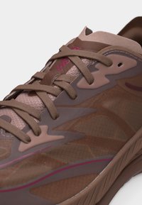 Κοντινή λήψη καφέ αθλητικού παπουτσιού με υφασμάτινο δίχτυ, συνθετικές επενδύσεις, χοντρές κορδόνια και μια μικρή ροζ λεπτομέρεια κοντά στη σόλα και τη γλώσσα.