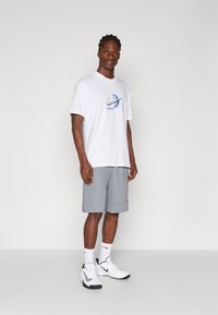 Λευκό κοντομάνικο μπλουζάκι με μπλε και γκρι λογότυπο Nike. Γκρι σορτς, λευκές κάλτσες και λευκά αθλητικά παπούτσια με μαύρες λεπτομέρειες.