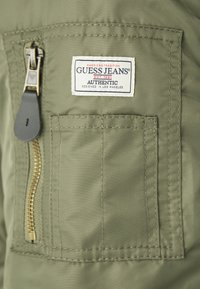 Zelená látková bunda s vreckom na zips so zlatými detailmi a bielou etiketikou s nápisom "GUESS JEANS AUTHENTIC, EST. 1981."