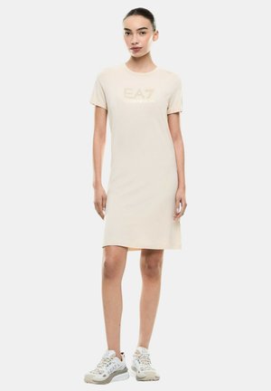 Donna che indossa un vestito beige a maniche corte fino al ginocchio con il logo "EA7 Emporio Armani", abbinato a sneakers bianche EA7 su sfondo bianco.