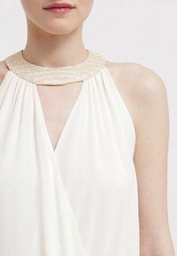 Robe blanche sans manches avec un col beige texturé, un tissu drapé avec un design enveloppant sur le devant, et une coupe en V.