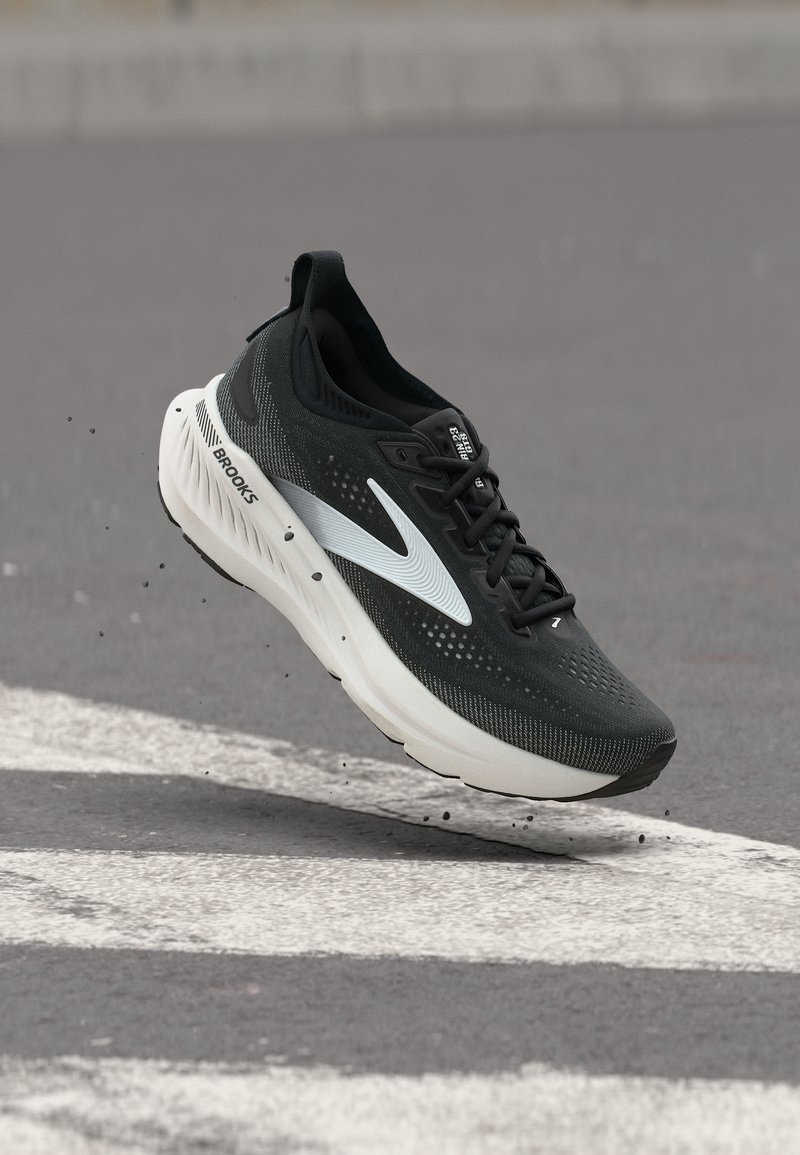 Chaussure de course Brooks noir et blanc en l'air au-dessus de l'asphalte avec des marquages blancs au sol, mettant en valeur la semelle et les détails du design latéral.