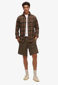 Camisa de franela a cuadros en verde oscuro y marrón, camiseta blanca, pantalones cortos oliva y zapatillas beige. Modelo de pie con las manos en los bolsillos.