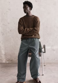 Hnedý rebrovaný sveter s voľným strihom, skombinovaný s širokými tehlovými kordrovými nohavicami. Pozadie tvorí neutrálny textúrovaný múr a stolička.