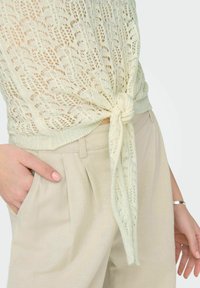 Top de punto crema claro con un patrón de encaje abierto, con un detalle de nudo en la cintura; combinado con pantalones beige con pliegues.