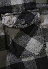 Juodos ir pilkos spalvos languotas marškinių audinys su matoma krūtinės kišene, kurioje yra sagos užsegimas ir tekstūruota, austinė medžiaga.