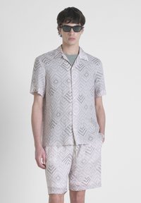 Camicia a maniche corte con bottoni e pantaloni corti abbinati in beige chiaro con un motivo geometrico. Il tessuto sembra leggero e traspirante.