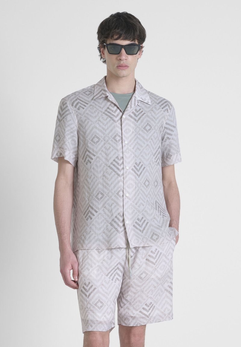 Camicia a maniche corte con bottoni e pantaloni corti abbinati in beige chiaro con un motivo geometrico. Il tessuto sembra leggero e traspirante.