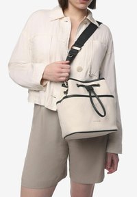 Bolso de lona tipo bucket en beige con acentos en negro. Cuenta con un cierre de cordón y una correa ajustable. La textura es suave con una forma estructurada.