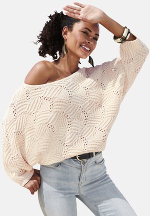 Lächelnde Frau mit lockigem Haar, die große Blatt-Ohrringe, einen lockeren, schulterfreien cremefarbenen Strickpullover, hellblaue Jeans und Armbänder trägt und die Augen abschirmt.