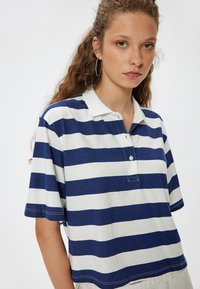 T-paita lyhyillä hihoilla, jossa on meri- ja valkoiset raidat, kaulus, nappilistat ja pehmeä pinta. Ominaisuuksiin kuuluu rento leikkaus ja sivusauman yksityiskohtia.