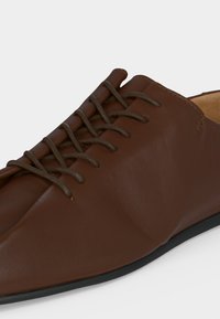 Chaussure habillée en cuir marron avec laçage fermé et semelle noire, montrant une texture détaillée et des coutures sur un fond clair.