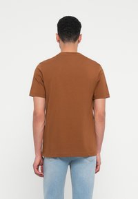 Καφέ κοντομάνικο T-shirt σε μαλακό, λείο ύφασμα, με κλασική στρογγυλή λαιμόκοψη και χαλαρή εφαρμογή, συνδυασμένο με ανοιχτό μπλε τζιν.