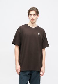 Ruda per didelę medvilninę marškinėlius su trumpomis rankovėmis ir baltu Adidas logotipu ant krūtinės, su lygaus paviršiaus tekstūra.