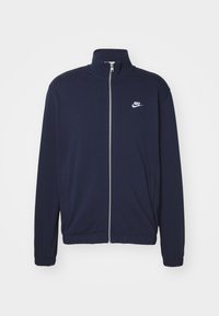 Granatowa kurtka na zamek z gładkiego materiału, z wysokim kołnierzem i elastycznymi mankietami. Małe białe logo Nike na górnej lewej stronie.