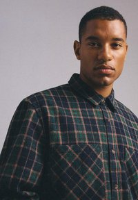 Czarna i zielona koszula w kratę z guzikami, z kołnierzykiem i dwiema przednimi kieszeniami. Materiał ma miękką, strukturalną powierzchnię.