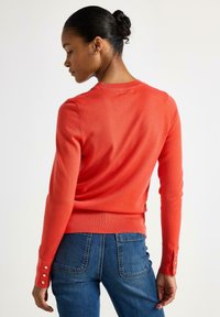 Rød tætsiddende sweater med rund halsudskæring, ribbet kant og dekorative knapdetaljer på ærmerne, parret med blå jeans.