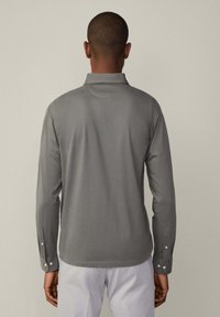 Muškarac u sivoj polo majici s dugim rukavima i bijelim hlačama, stoji okrenut leđima prema kameri na jednoličnoj pozadini.