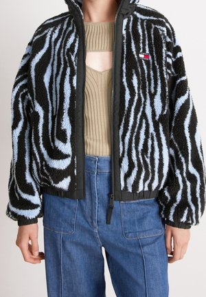 Persona que lleva una chaqueta de forro polar con rayas de cebra en negro y azul claro, un top beige acanalado con abertura y vaqueros azules de cintura alta.