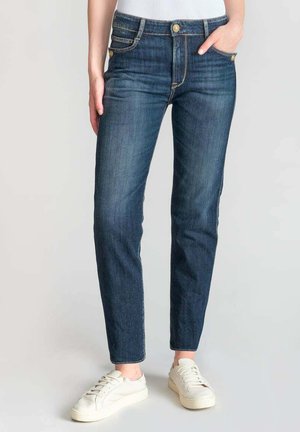 Dunkelblaue Jeans aus Denim mit einer taillierten Passform, ausgestattet mit einer Fünf-Pocket-Optik, einem Reißverschluss und einem leicht zulaufenden Bein. Getragen mit weißen Sneakers.