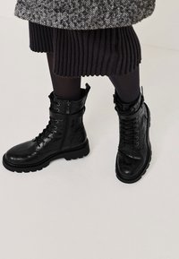Bottes de combat noires à lacets avec fermeture éclaire sur le côté, portées avec des collants noirs et une jupe noire côtelée sous un manteau en chevron.