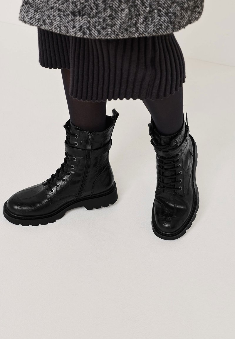 Bottes de combat noires à lacets avec fermeture éclaire sur le côté, portées avec des collants noirs et une jupe noire côtelée sous un manteau en chevron.