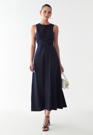 Robe navy avec un buste ajusté sans manches et une taille drapée. Se prolonge jusqu'à un ourlet mi-genou. Associée à un sac à main blanc et des talons noirs à lanières.