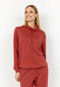 Rode pullover met een col, lange mouwen en een relaxed fit. Bevat trekkoorden en de tekst "AVONTUREN BEGINNEN" gedrukt op de voorkant.