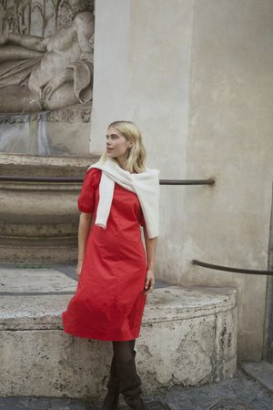 Punainen puuvillamekko, jossa on pullistuvat hihat ja polven pituinen helma, yhdistetty valkoiseen neulottuun villapaitaan, joka on asetettu harteille. Ruskeat polven korkeudelle ulottuvat saappaat.