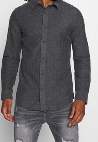 Camisa de botón de color gris oscuro hecha de una tela suave, con un cuello estructurado, mangas largas y detalles de costura limpios. Combinada con jeans desgastados.
