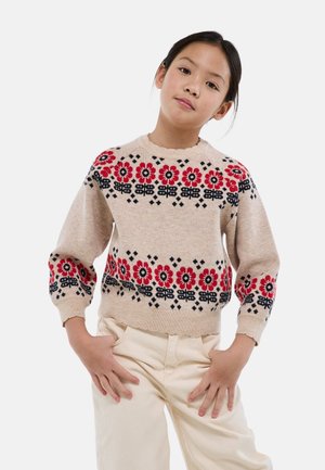 Jeune fille debout, les mains dans les poches d'un pantalon crème, portant un pull beige avec un motif floral rouge et noir sur la poitrine et les manches.