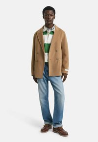 Camelkleurige double-breasted jas met zakken, gelaagd over een groen-wit gestreept polo-shirt, in combinatie met lichtblauwe jeans en bruine schoenen.